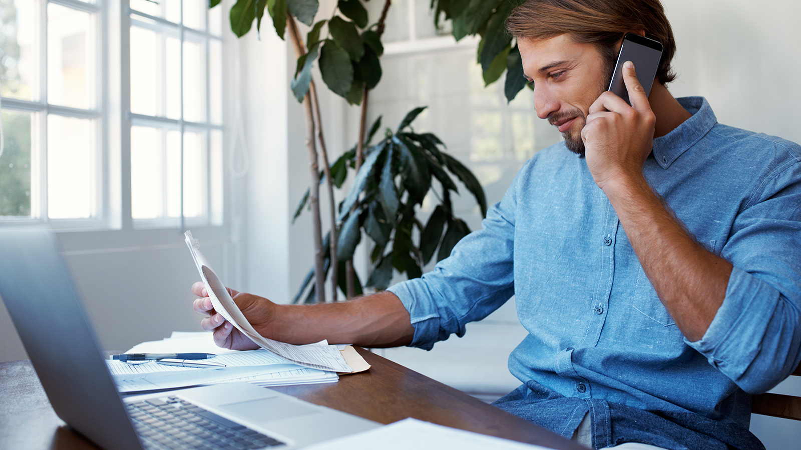 Man seated at desk with laptop reviewing paperwork.