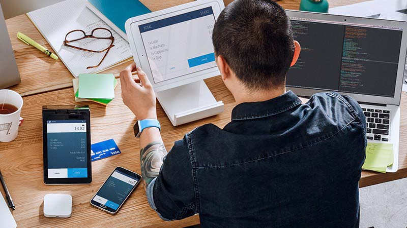 Man with a laptop, tablet, mobile phone, papers and Square payment device on a desk in front of him.